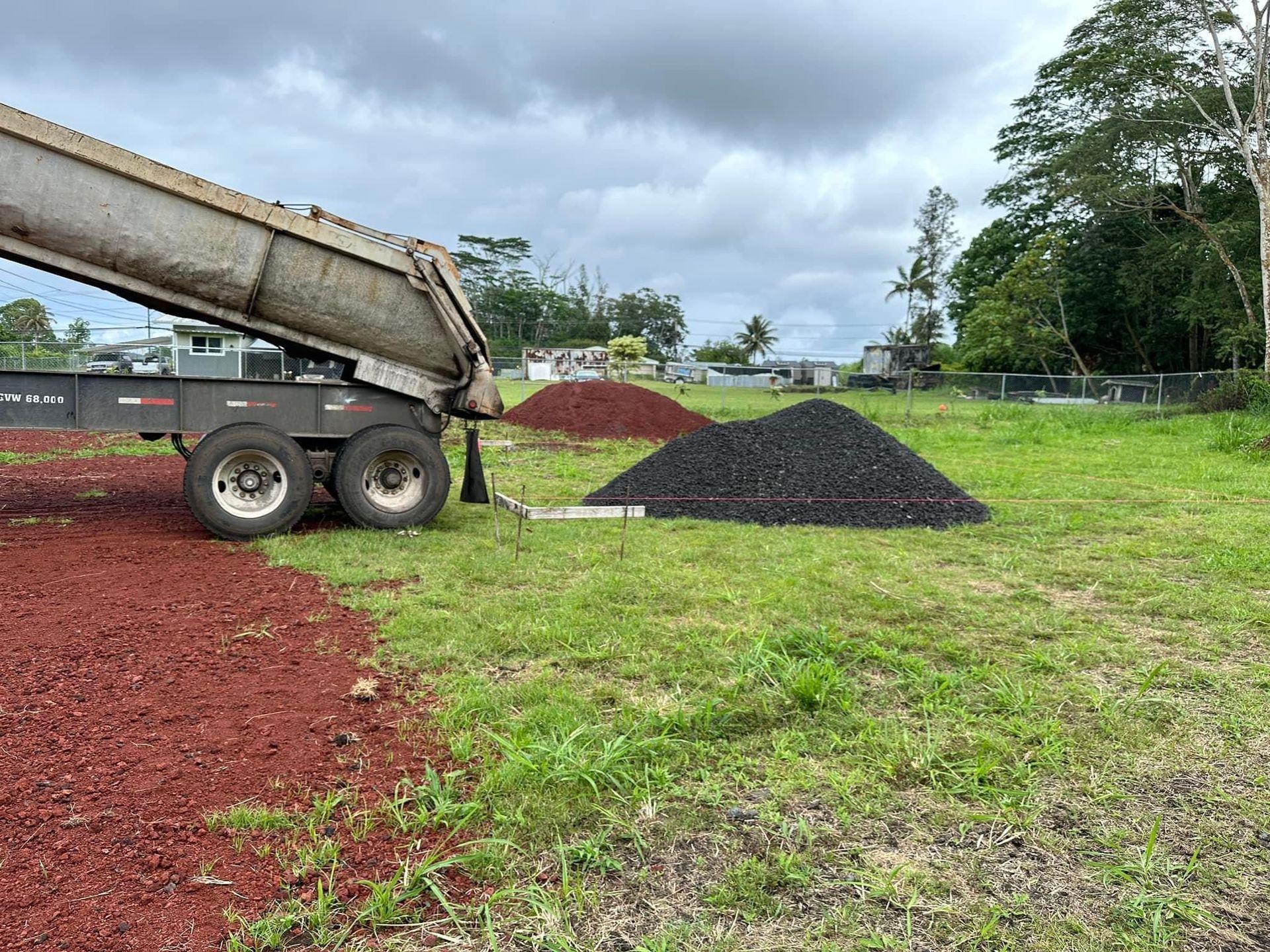 Dump truck unloading red soil, beside piles of black gravel on a grassy field.