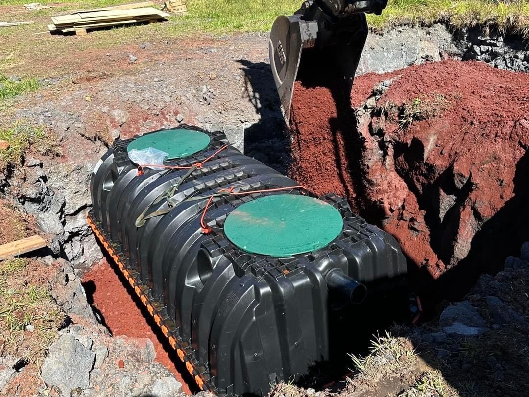 Excavator placing a black septic tank into a dug-out hole surrounded by dirt and grass.