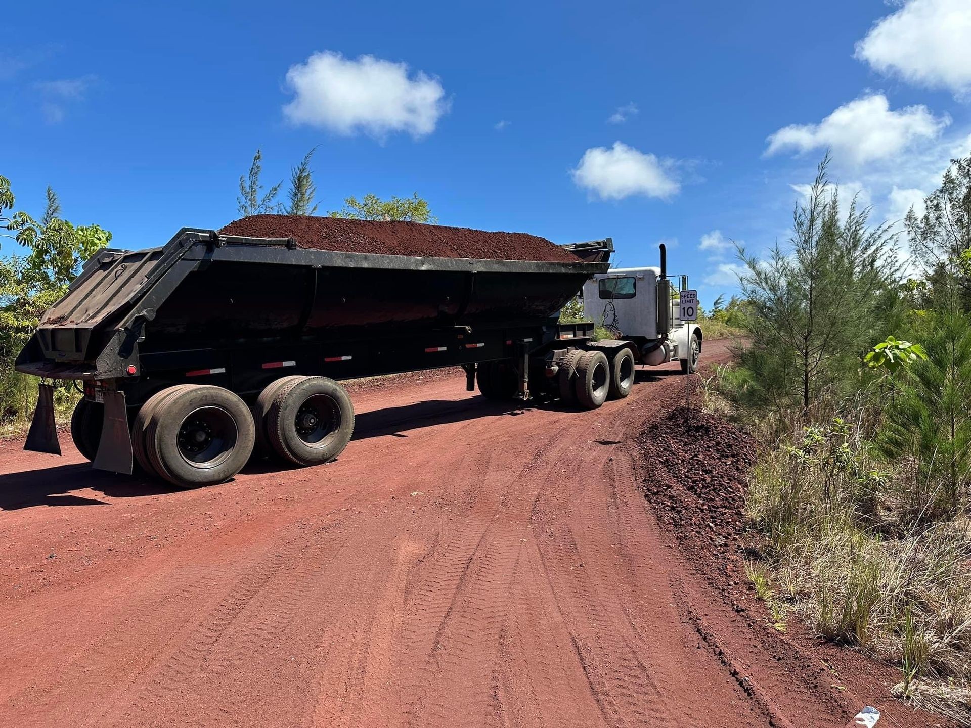 Dump truck loaded with red dirt on a rural red dirt road under a clear blue sky.