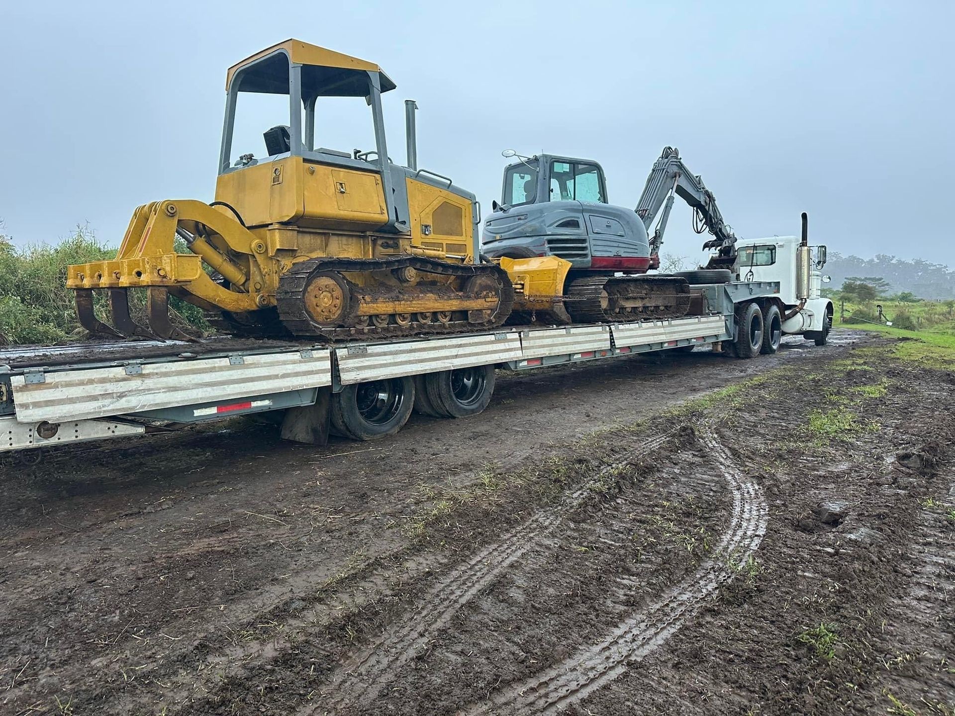 Bulldozer and excavator loaded on a flatbed truck on a muddy road.