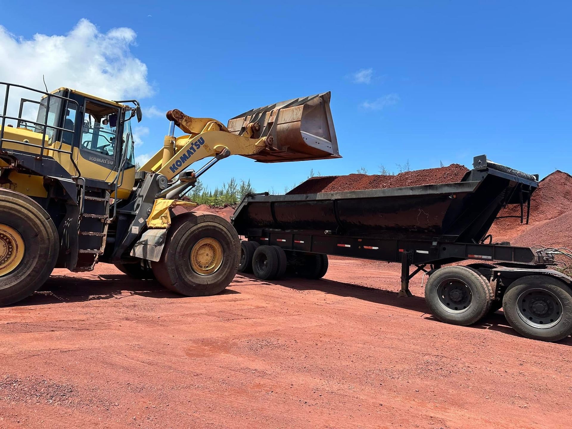 Large yellow bulldozer loading red soil into an open trailer on a bright sunny day.