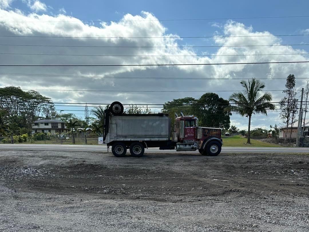 Dump truck parked on a gravel road with lush tropical vegetation and power lines in the background.